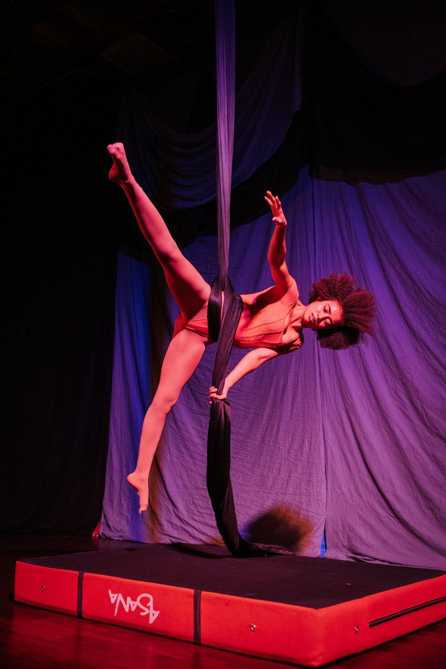 Aerial performer in red outfit gracefully balances on a pole under dramatic stage lighting.