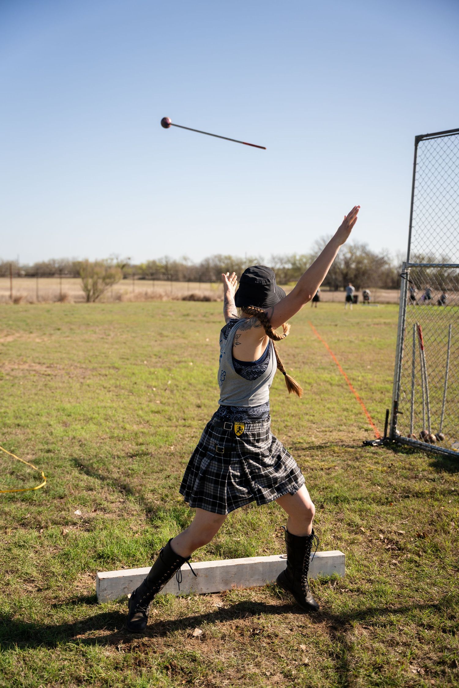 Woman in plaid skirt and boots throwing a hammer at a Highland Games competition outdoors.