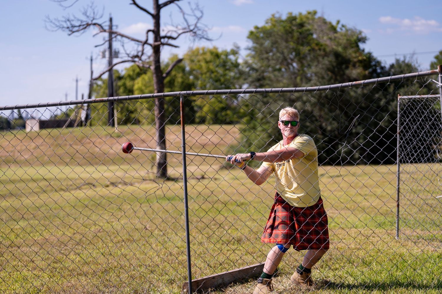 Person wearing a kilt practices baseball swings in a sunny field with chain link fence and trees in background.