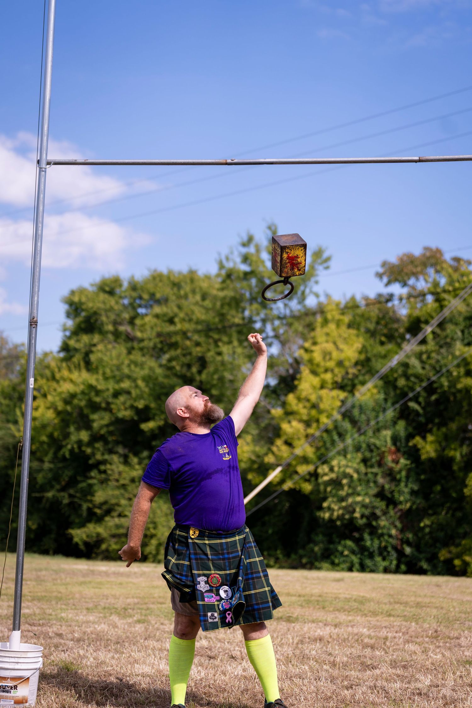 Athlete in purple shirt and kilt performs weight throw technique in three sequential action shots on grassy field.