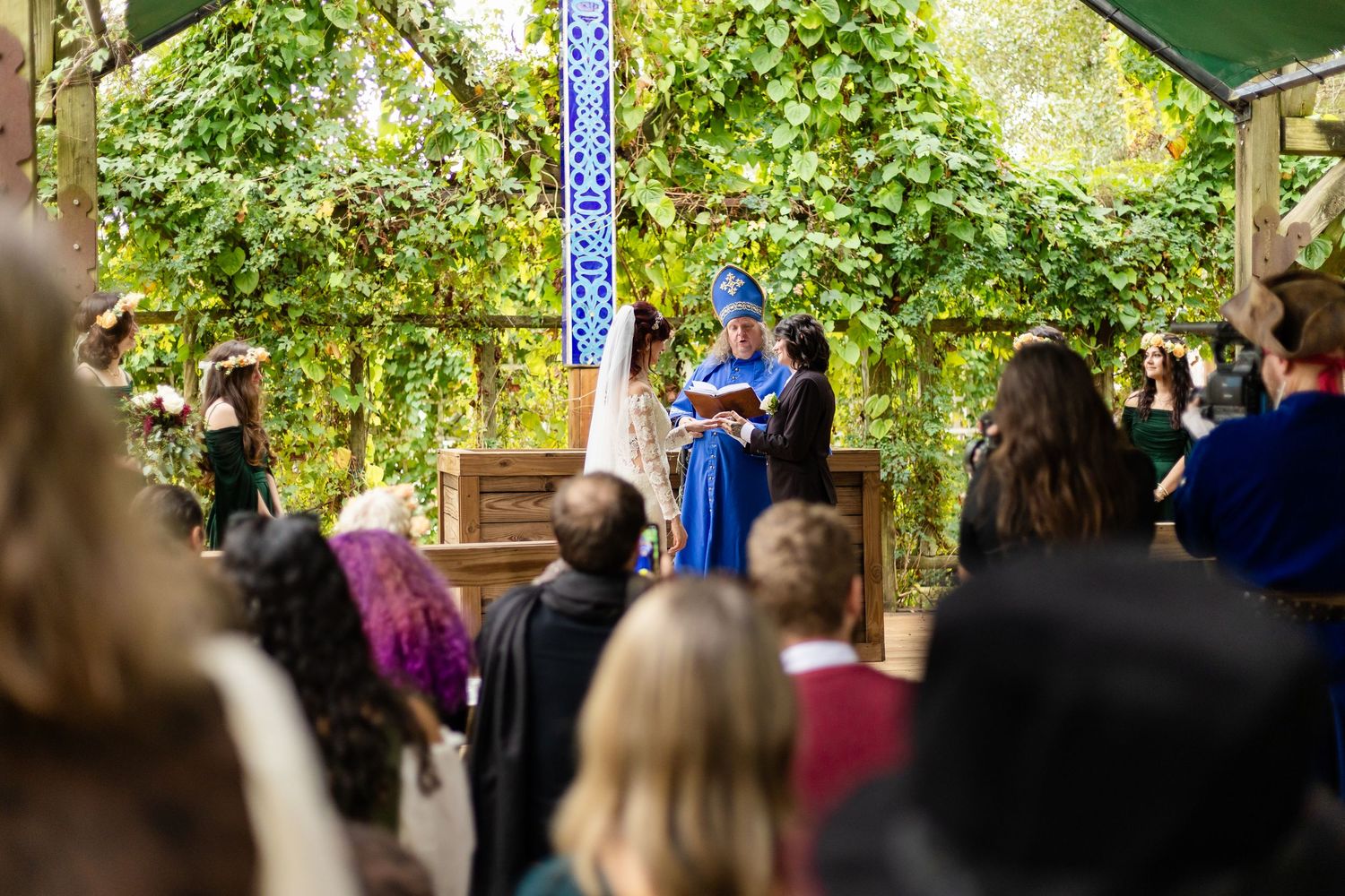 Wedding ceremony takes place in a lush outdoor garden setting with guests seated under green foliage.