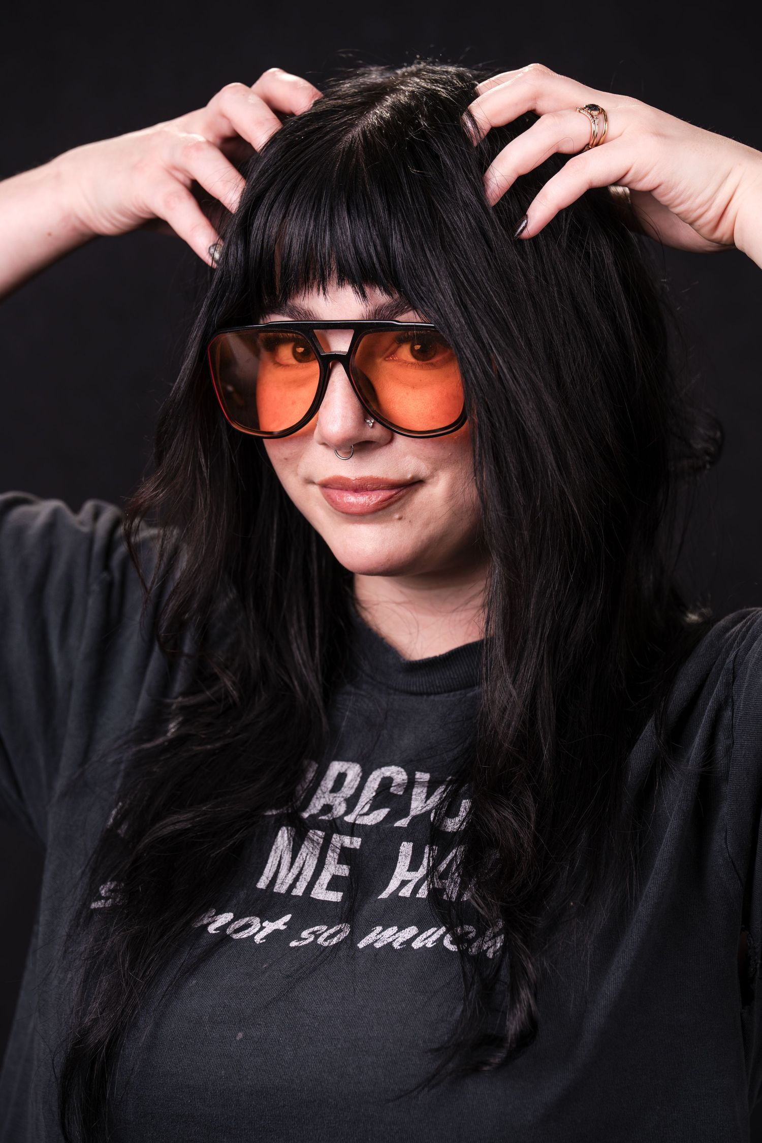 A person with dark hair and sunglasses poses playfully against a black background while wearing a black t-shirt.