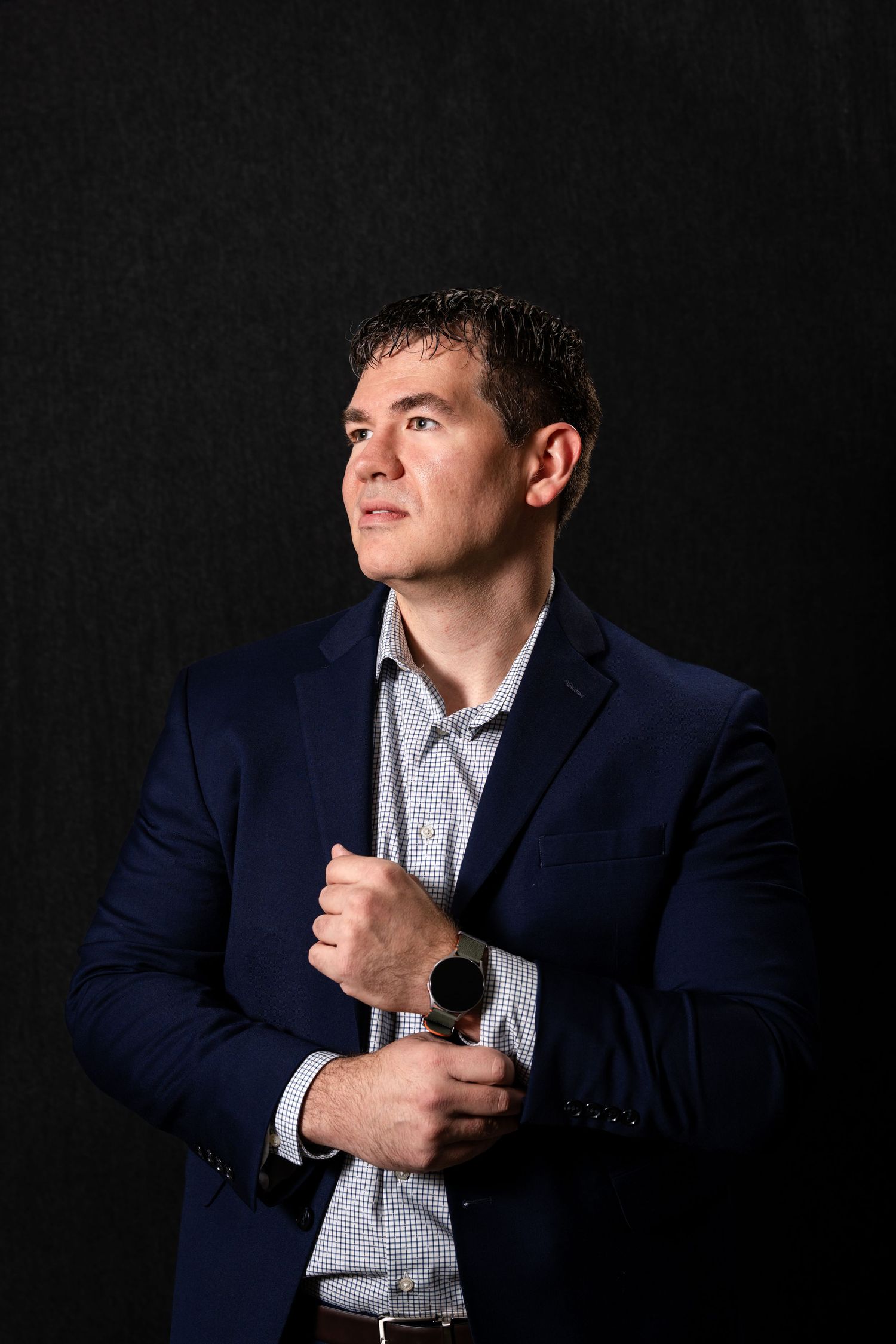 Professional studio portrait of someone in a navy suit and patterned dress shirt against a dark background.