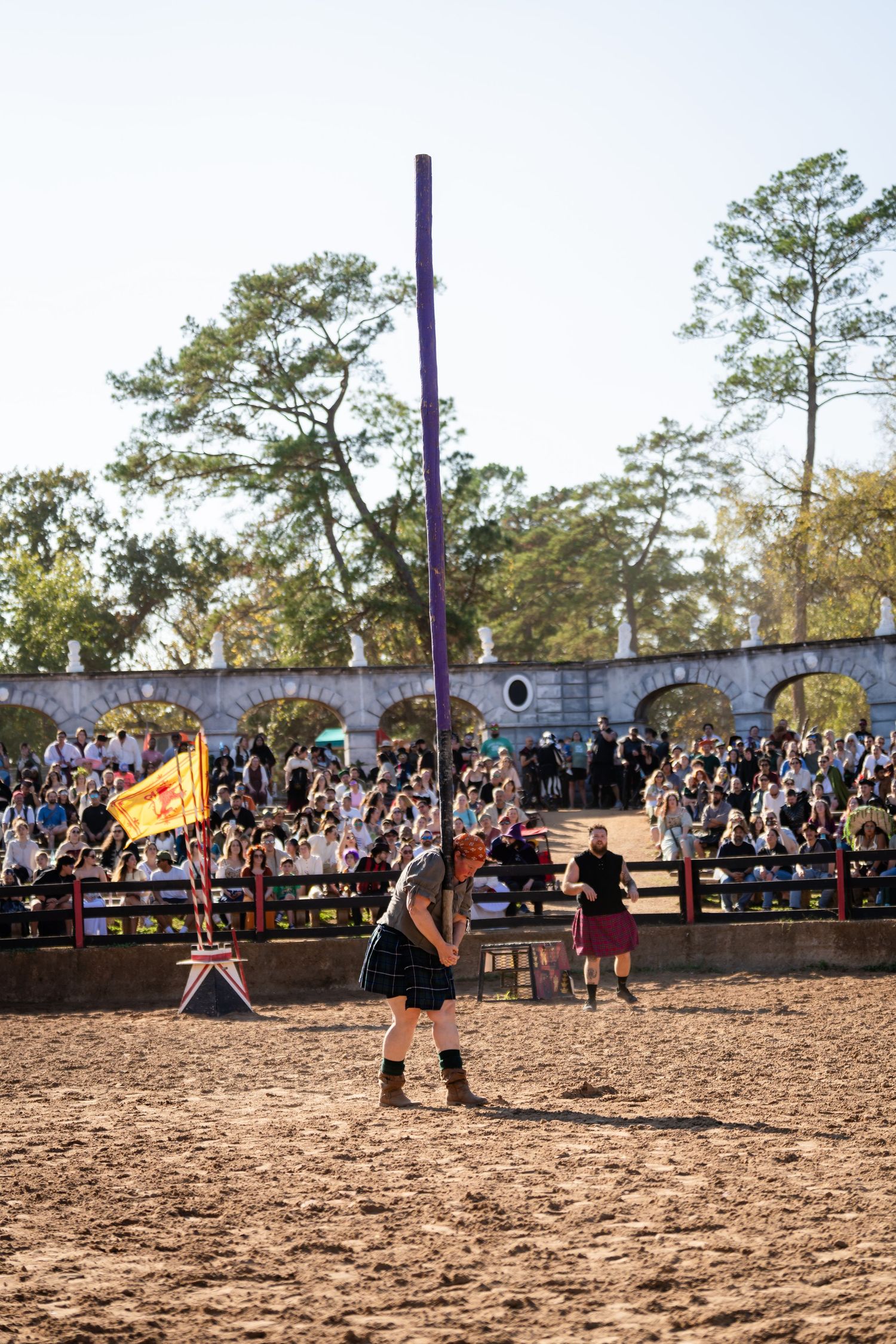 Medieval knights duel with swords in a dirt arena as spectators watch from bleachers at a Renaissance festival.