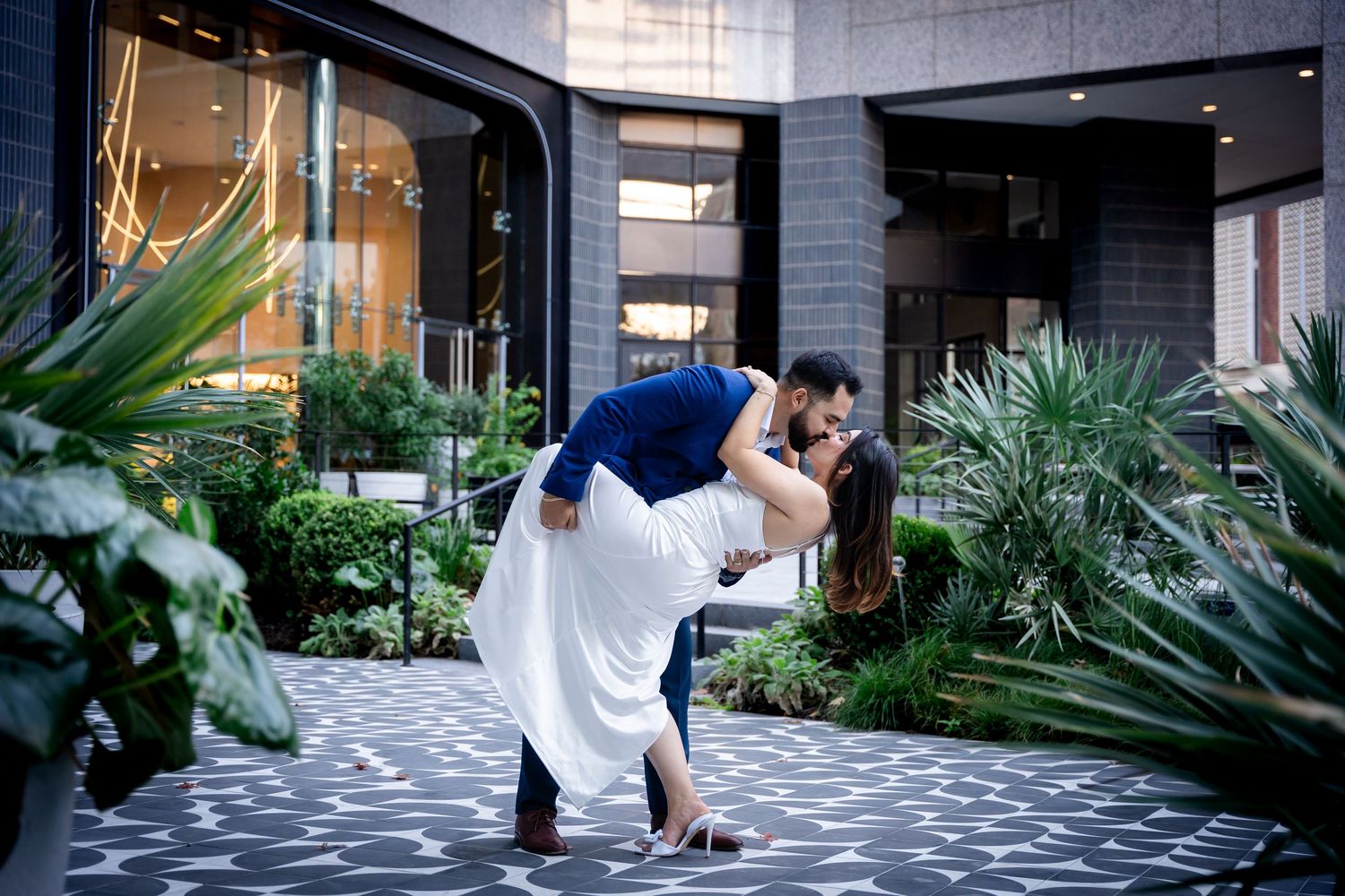 A couple shares a romantic moment in a modern indoor garden space with geometric floor patterns and tropical plants.