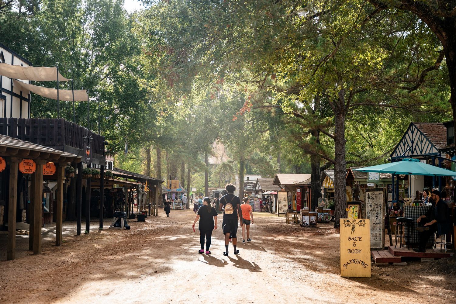 Tree-lined path in a park with people walking and outdoor dining areas visible in the distance.