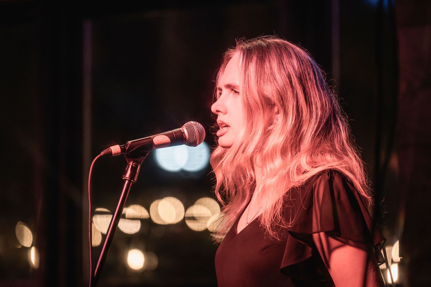 Singer with pink-tinted hair performs on stage with microphone under dramatic lighting at a concert venue.
