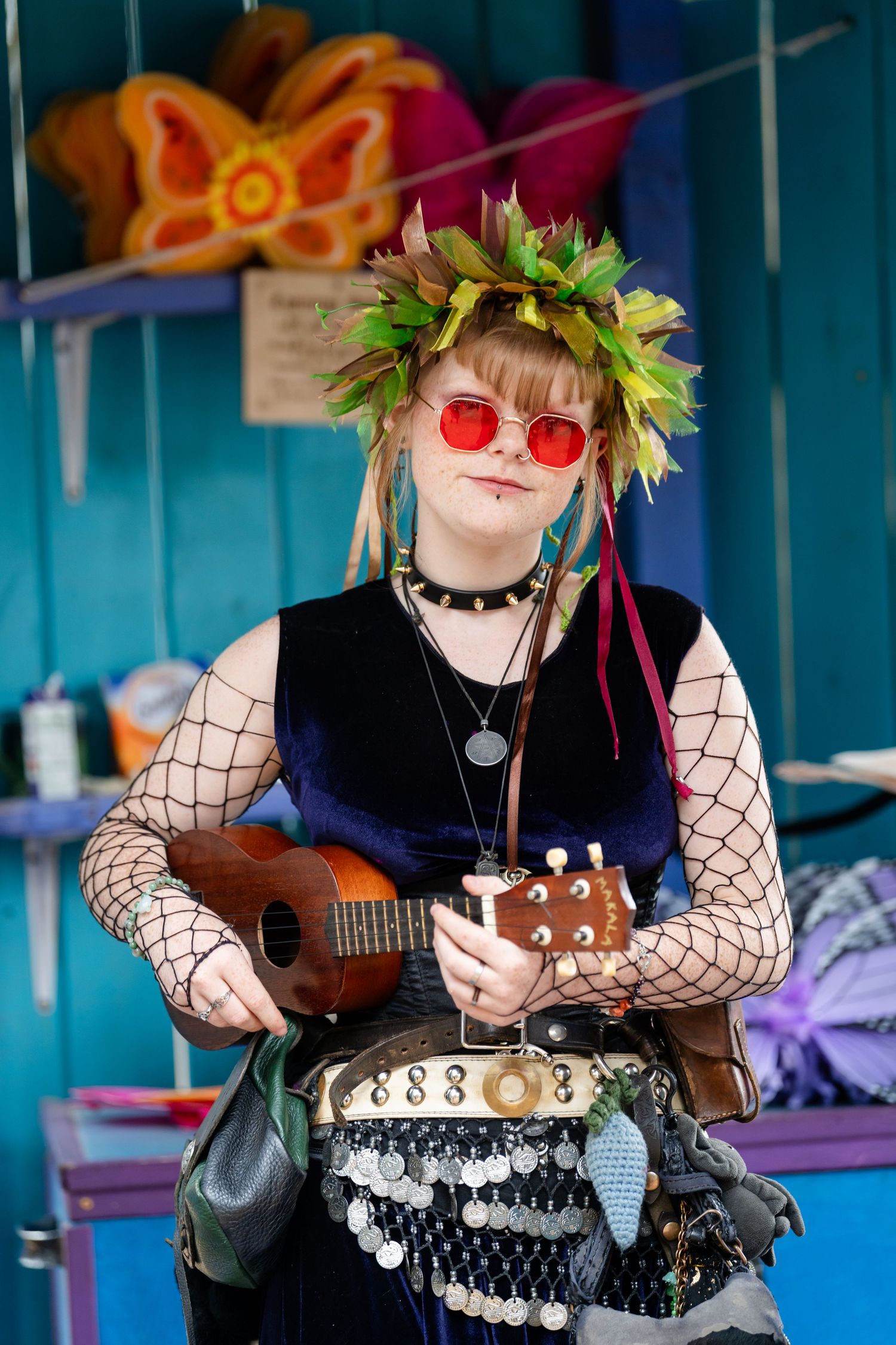 Musician in fishnet sleeves and red sunglasses plays ukulele while wearing green leaf crown against blue background.