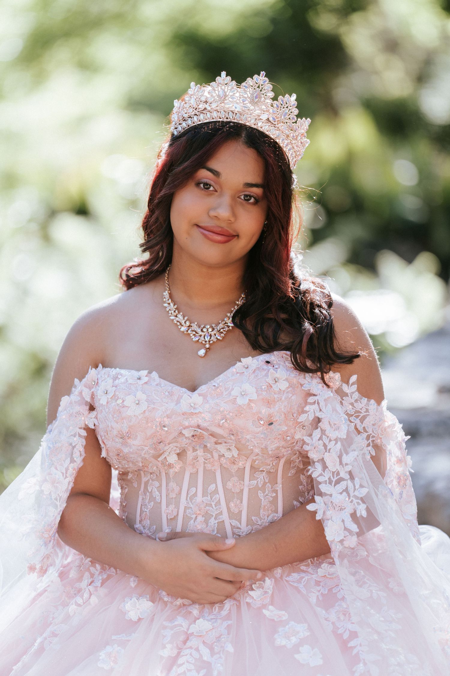 Young woman in a blush pink off-shoulder quinceañera ball gown with floral lace, crystal crown, and diamond necklace outdoors.