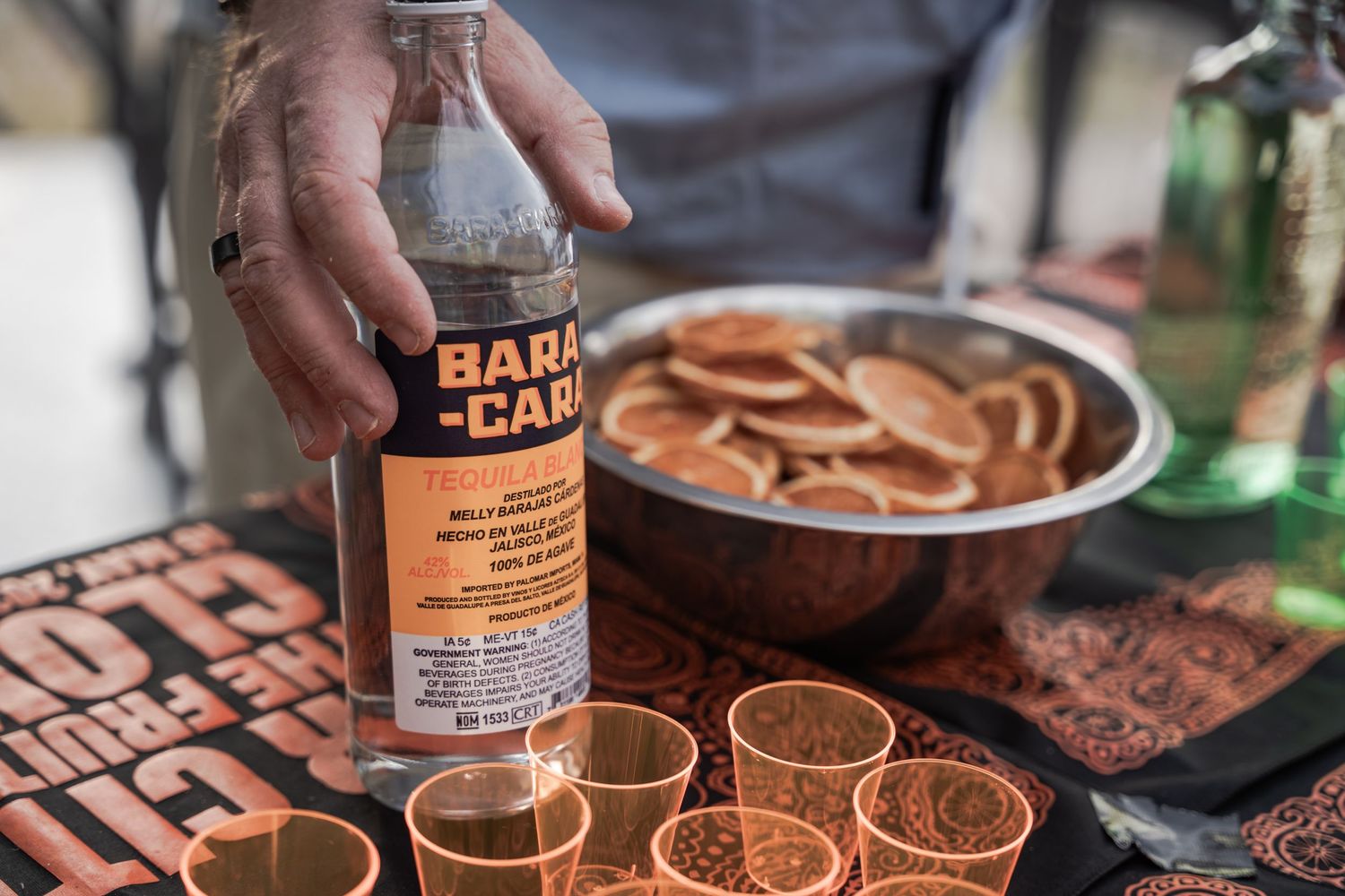 Hand pouring Bara Cara tequila into copper shot glasses beside a bowl of sliced oranges on a bar table.