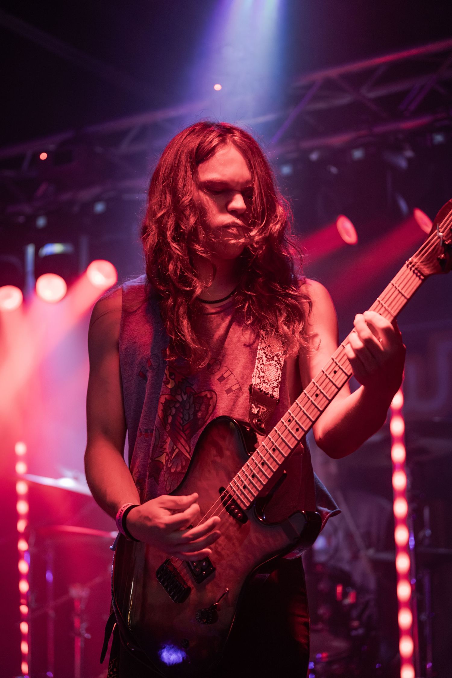 Long-haired guitarist performing on stage under dramatic red and purple concert lighting.