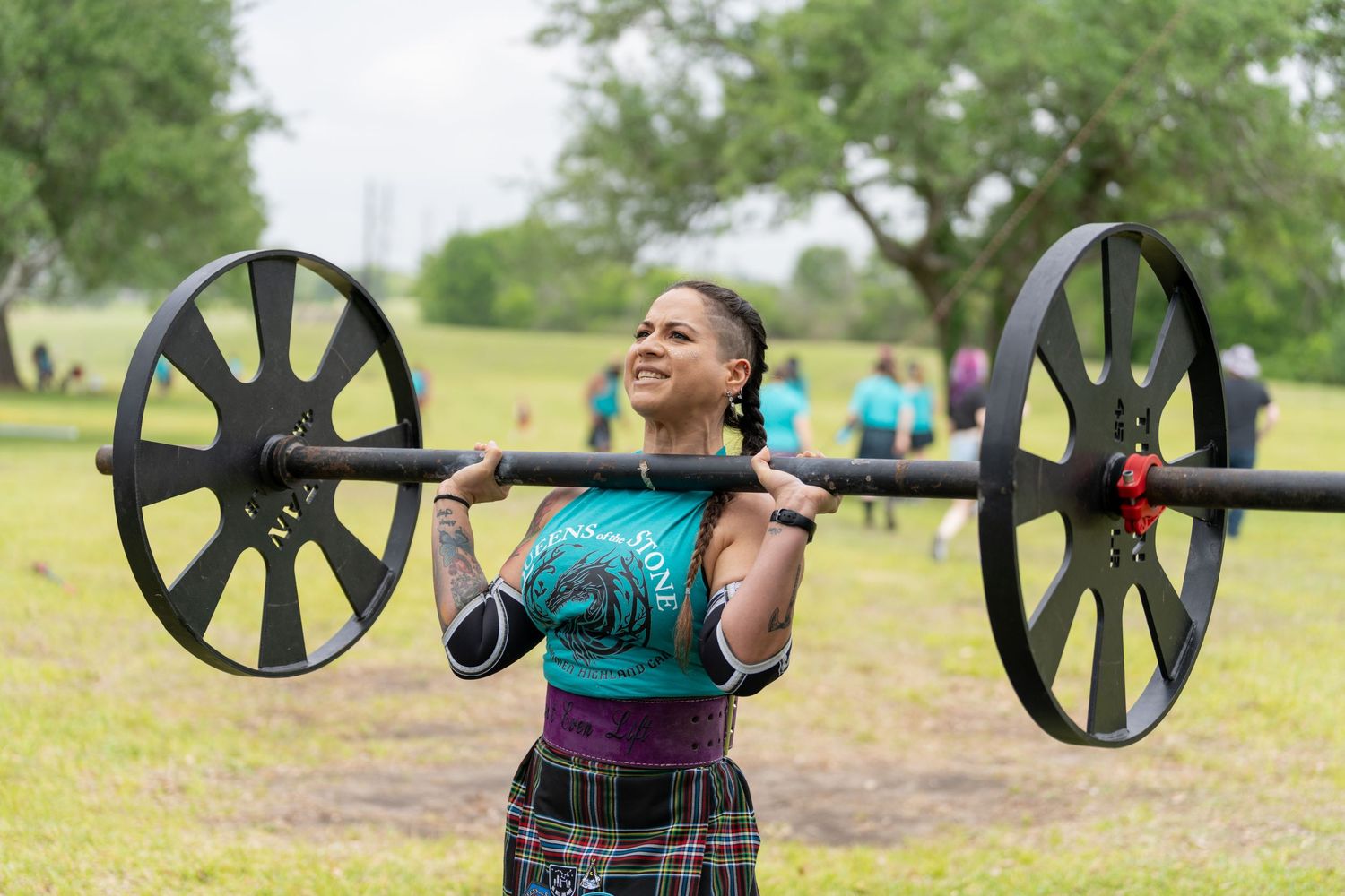 Woman in tartan skirt lifting heavy barbell outdoors at Highland Games fitness competition.