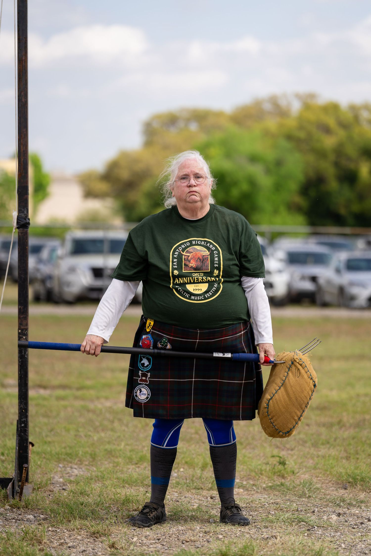 Person in green anniversary t-shirt and tartan kilt holding a basket, standing at Highland Games field.
