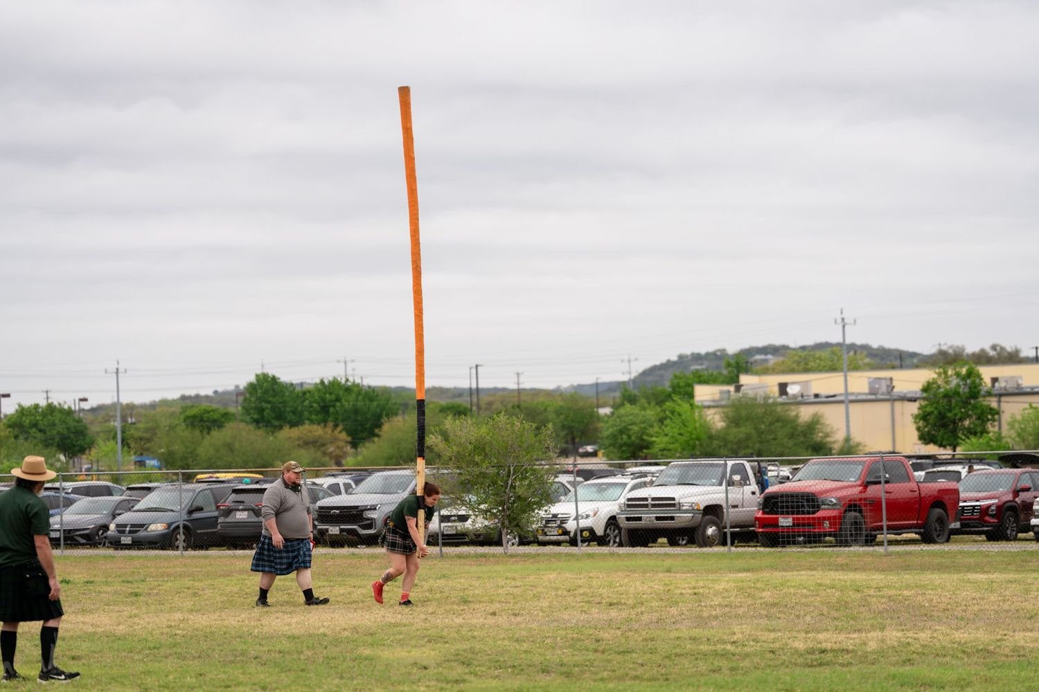People playing outdoor games on a green field with parked cars and rolling hills in the background under cloudy skies.