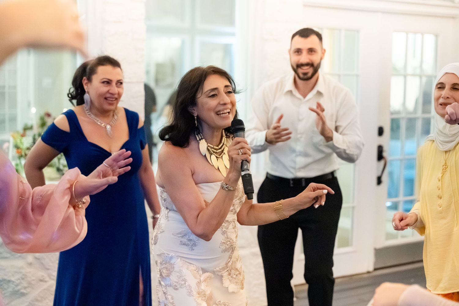 Woman singing with microphone at wedding reception, guests clapping and celebrating in elegant venue.