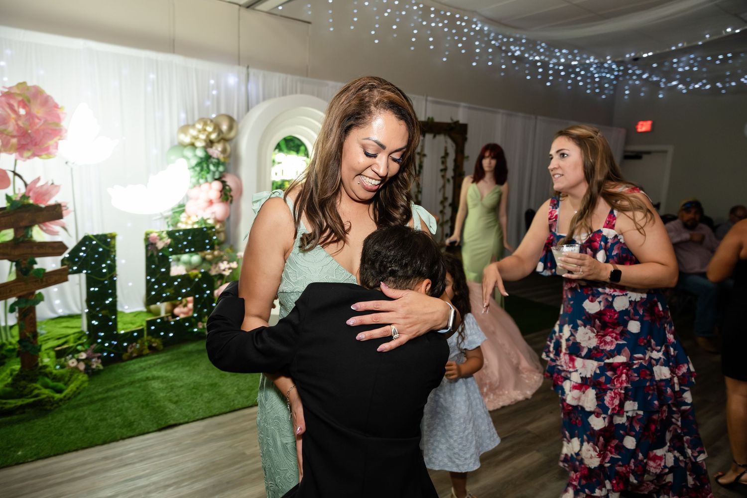 Woman hugging a child at a quinceañera celebration with decorative floral backdrop and number 15 display.