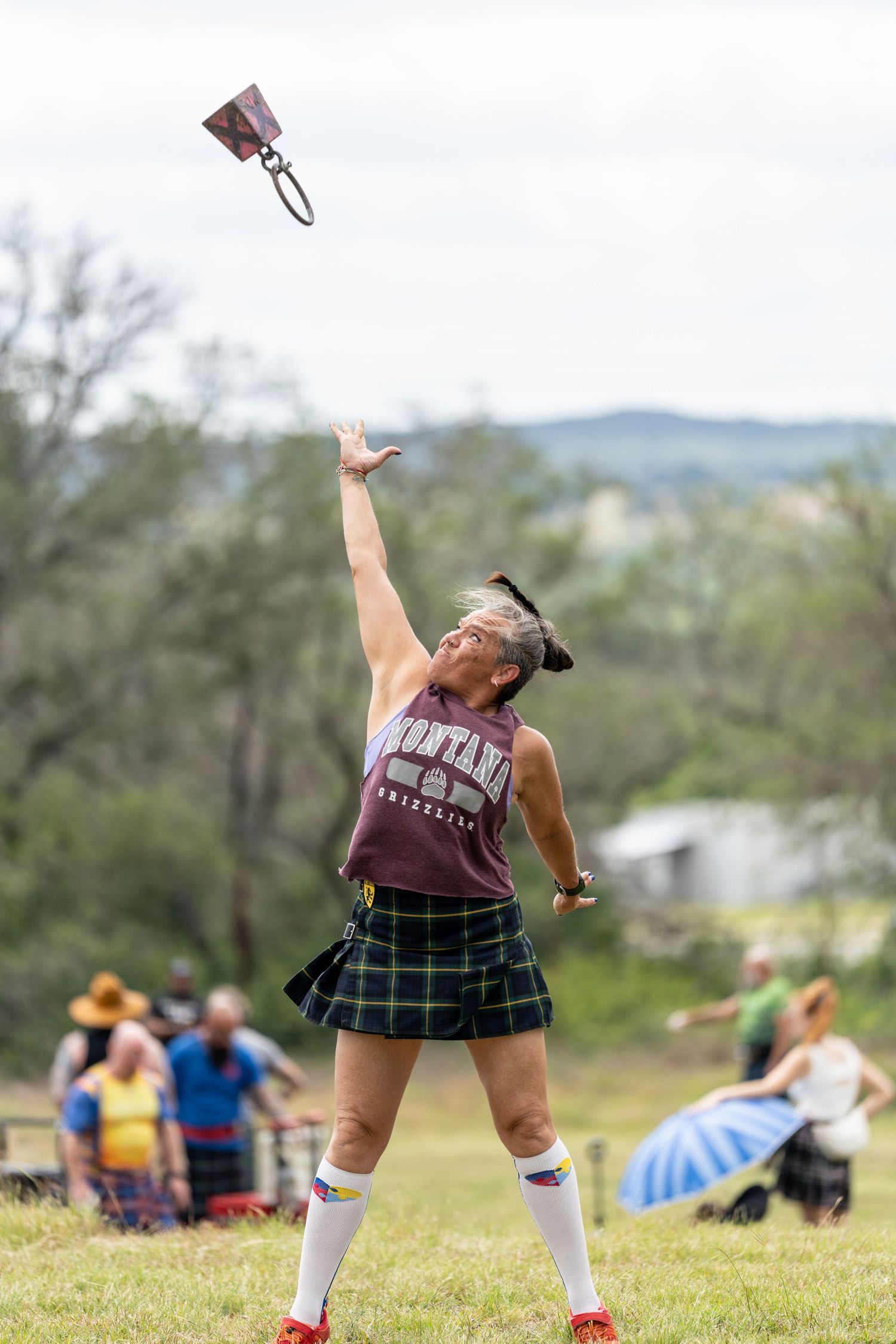 Woman in tartan kilt throws weight over bar at Highland Games competition outdoors.