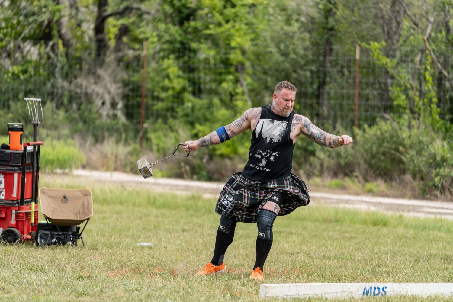 Tattooed man in kilt competes in Highland Games throwing event on grassy field.