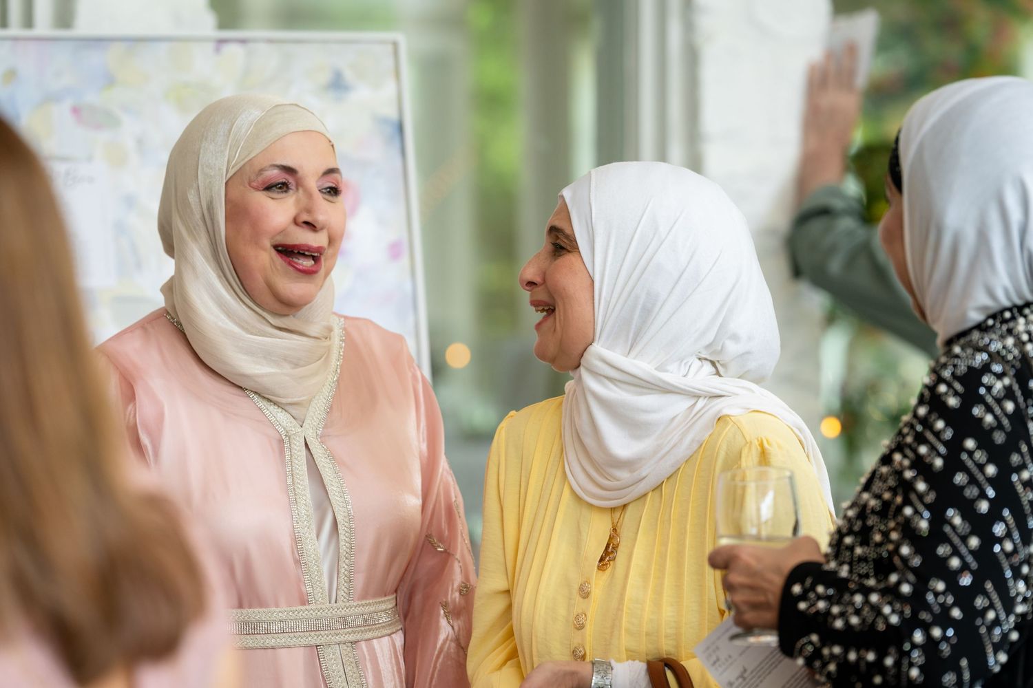 Two women wearing hijabs smile and chat at a social gathering, one in pink and one in yellow.
