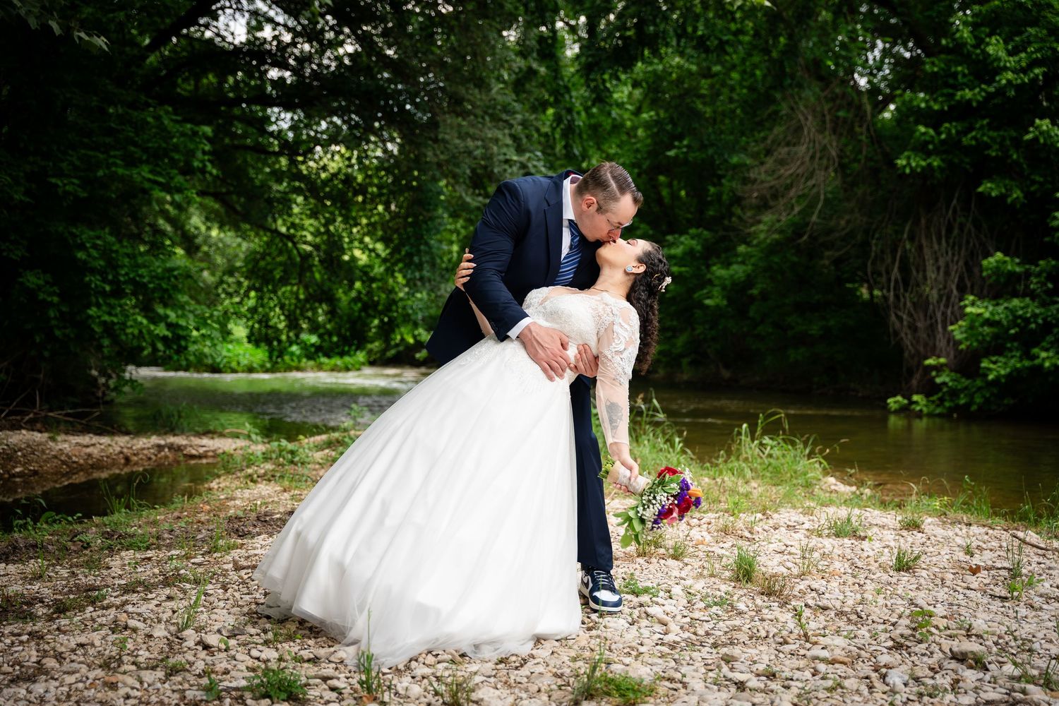 A groom dips his bride for a romantic kiss near a serene creek surrounded by lush green trees on their wedding day.