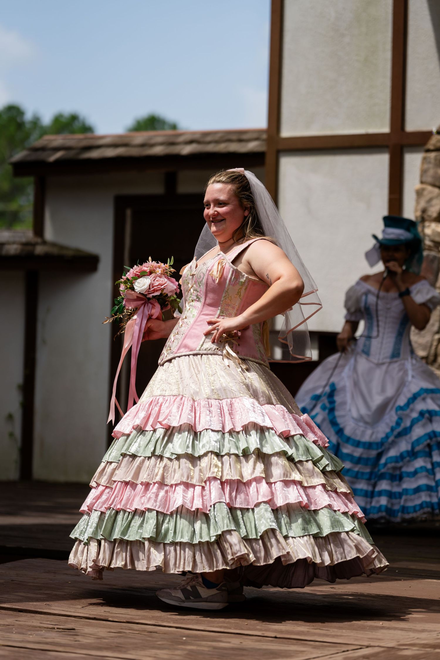 Woman in pink ruffled bridal gown holding bouquet twirling on outdoor stage at Renaissance faire.
