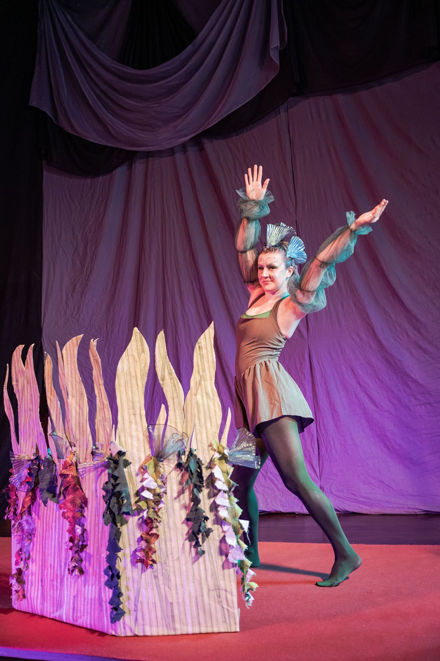 A dancer in a gold costume with teal accessories performs on stage beside a colorful floral prop under purple lighting.