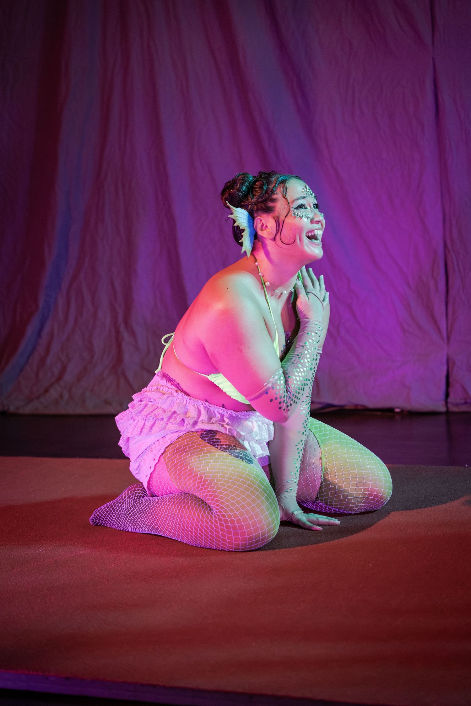 Performer in pink ruffled skirt and fishnet tights kneeling on stage under colorful lighting.