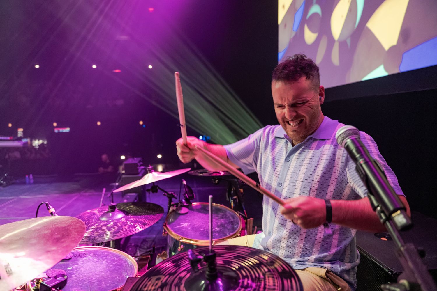 Energetic drummer playing cymbals on stage with purple concert lighting and projection screen backdrop.