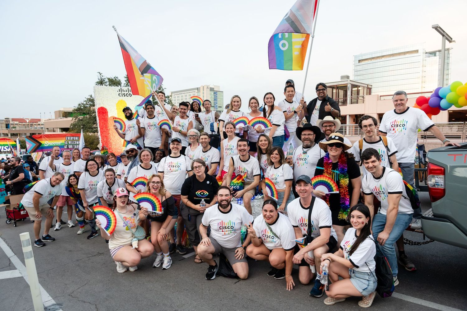 Large group in white tees celebrating Pride parade, holding rainbow flags outdoors at sunset.