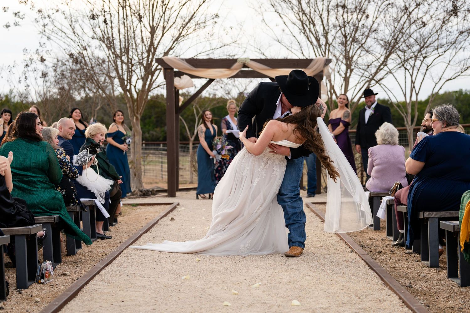 Groom in cowboy hat dips bride in white gown during outdoor wedding ceremony with seated guests lining the aisle.