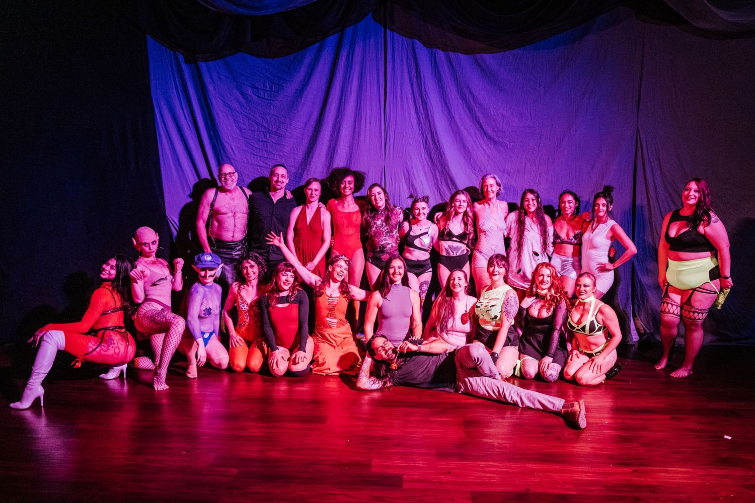 Large group of burlesque performers posing together on stage under pink and blue lights after a show.