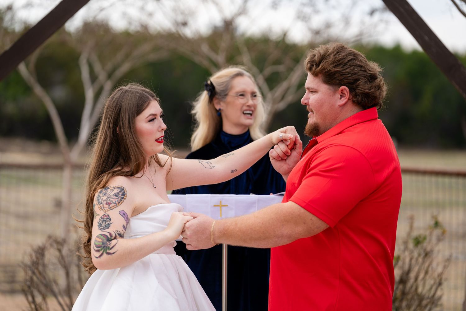 Outdoor wedding ceremony with tattooed bride, groom in red polo, and officiant holding a cross book.