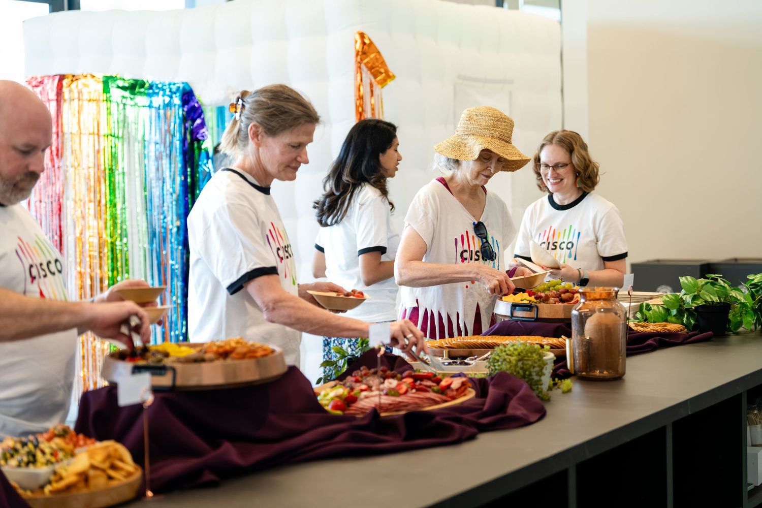 People in white t-shirts serving food at a colorful buffet table decorated with rainbow streamers.