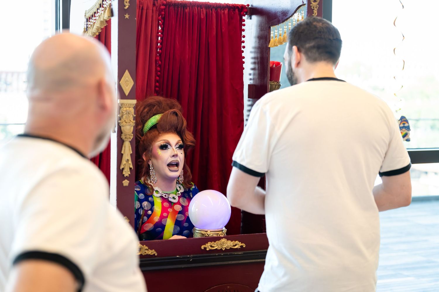Drag queen fortune teller in colorful costume performs for guests at a carnival booth event.