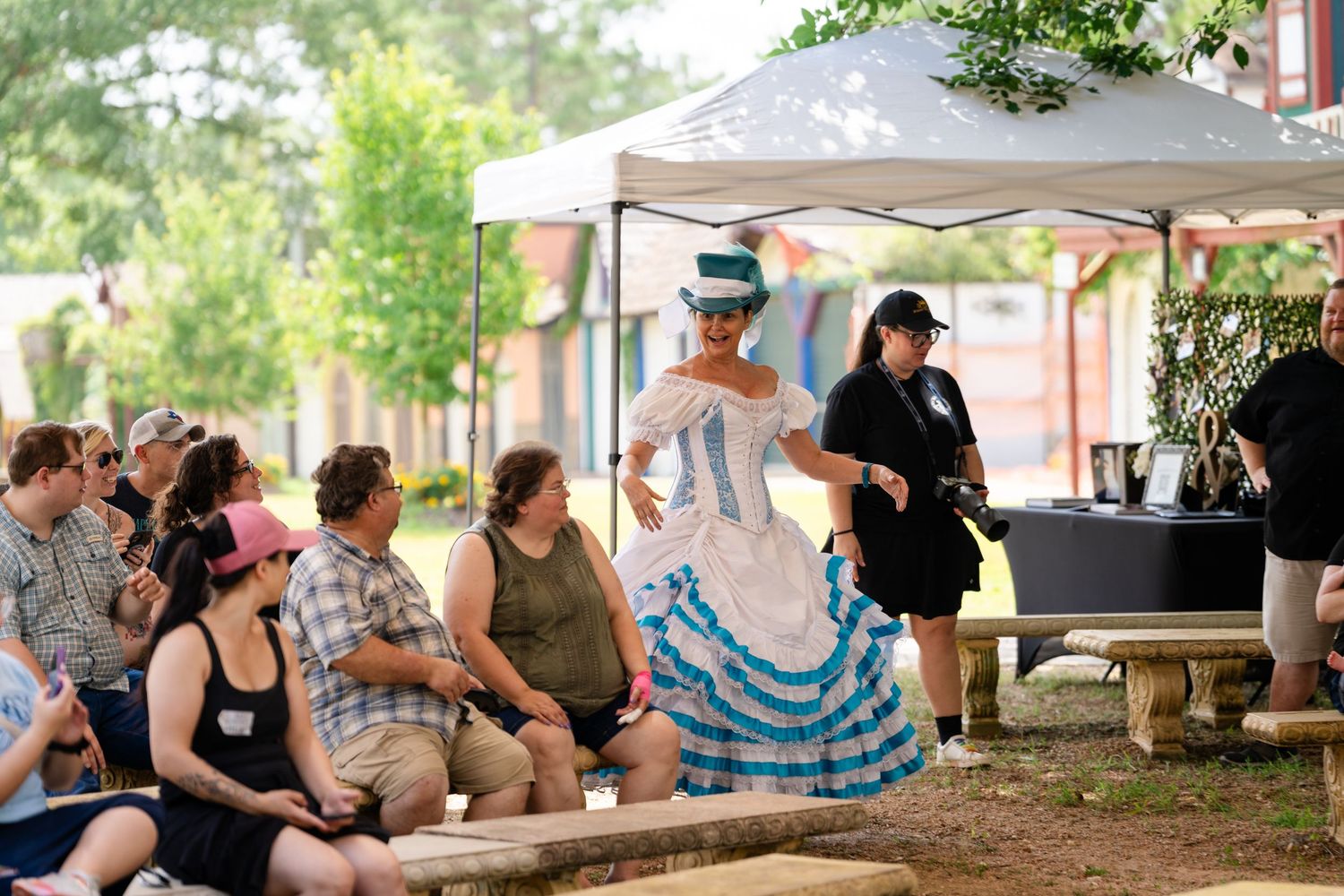 Woman in Victorian blue and white dress performs at outdoor historical costume event with spectators watching nearby.