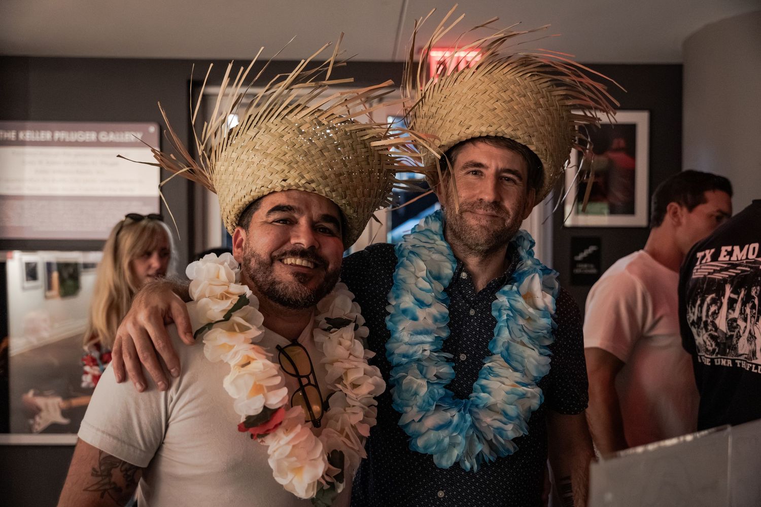 Two men wearing straw hats and tropical leis smile together at a Hawaiian-themed party indoors.