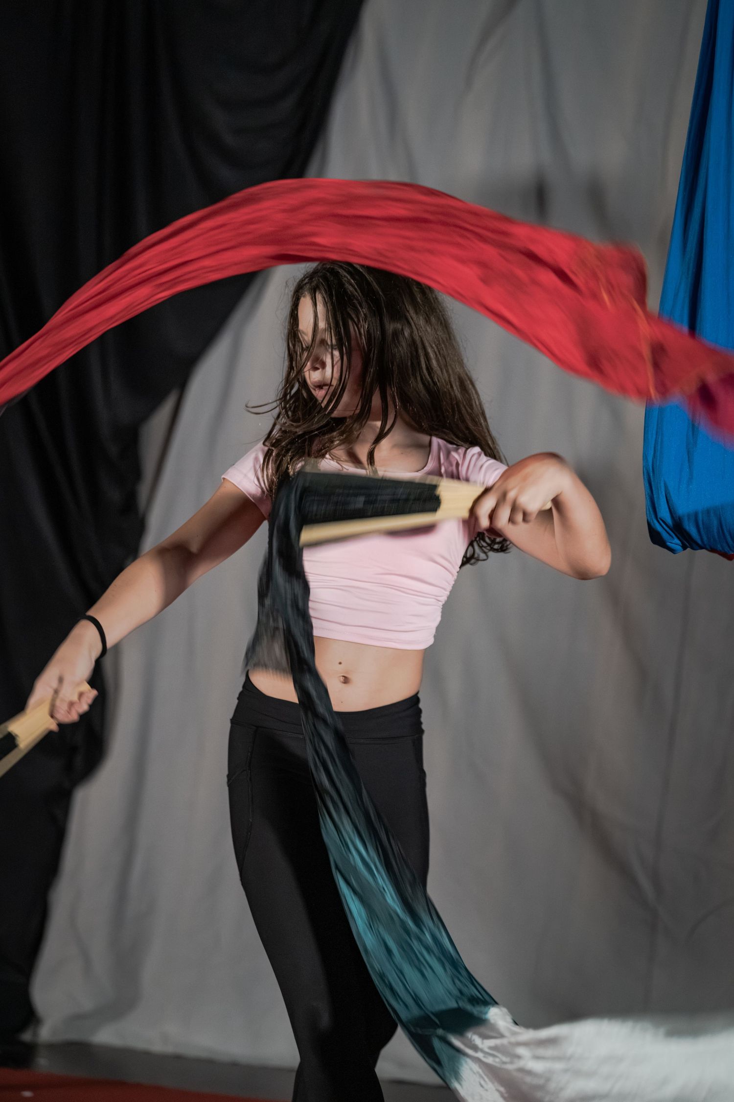 Young woman performing dynamic flag dancing with colorful red and teal silk flags in a studio setting.