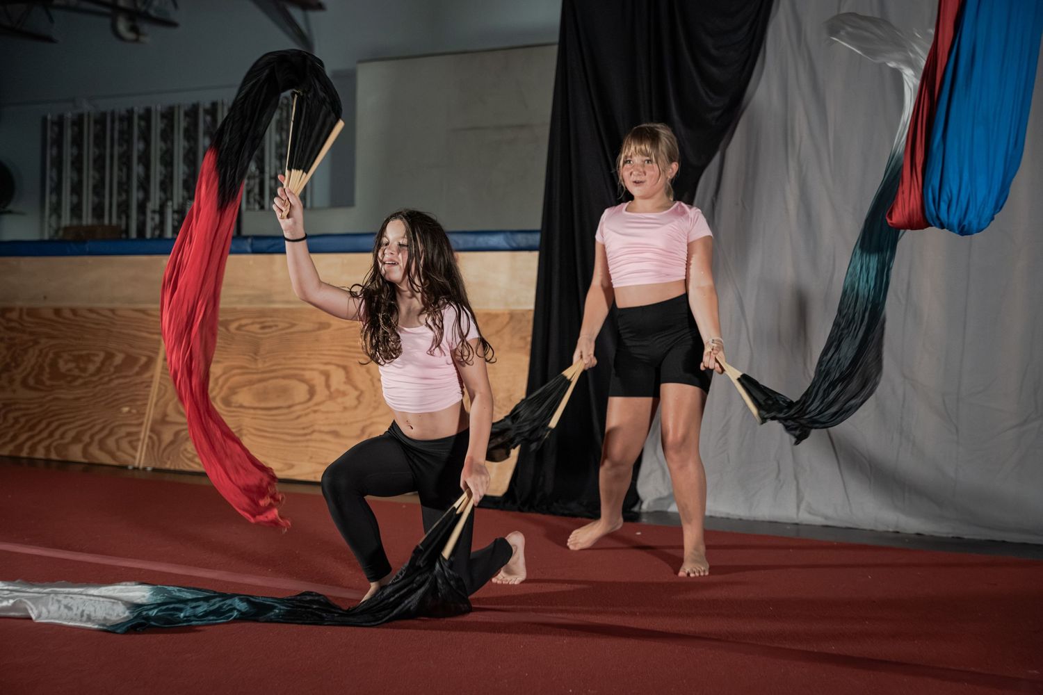 Two girls in pink tops practice rhythmic gymnastics with colorful ribbons in an indoor gymnasium.