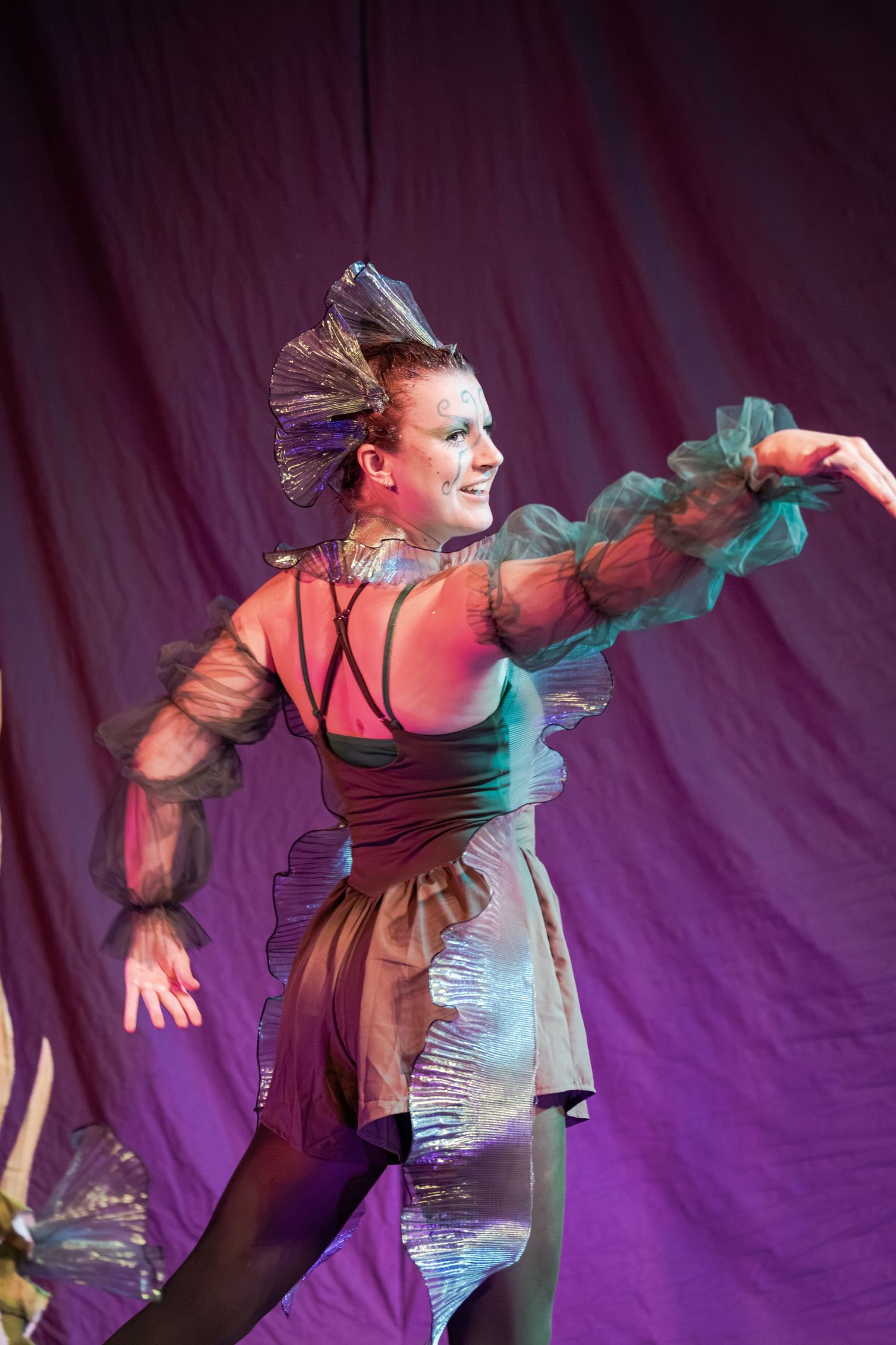 A female dancer performs on stage in an iridescent costume with sheer sleeves and an ornate headpiece against a purple backdrop.