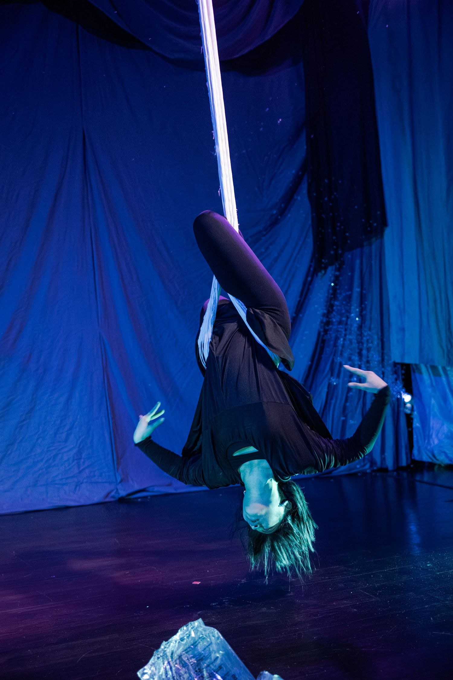 Aerial silk performer in black outfit hanging upside down on stage with blue dramatic lighting.