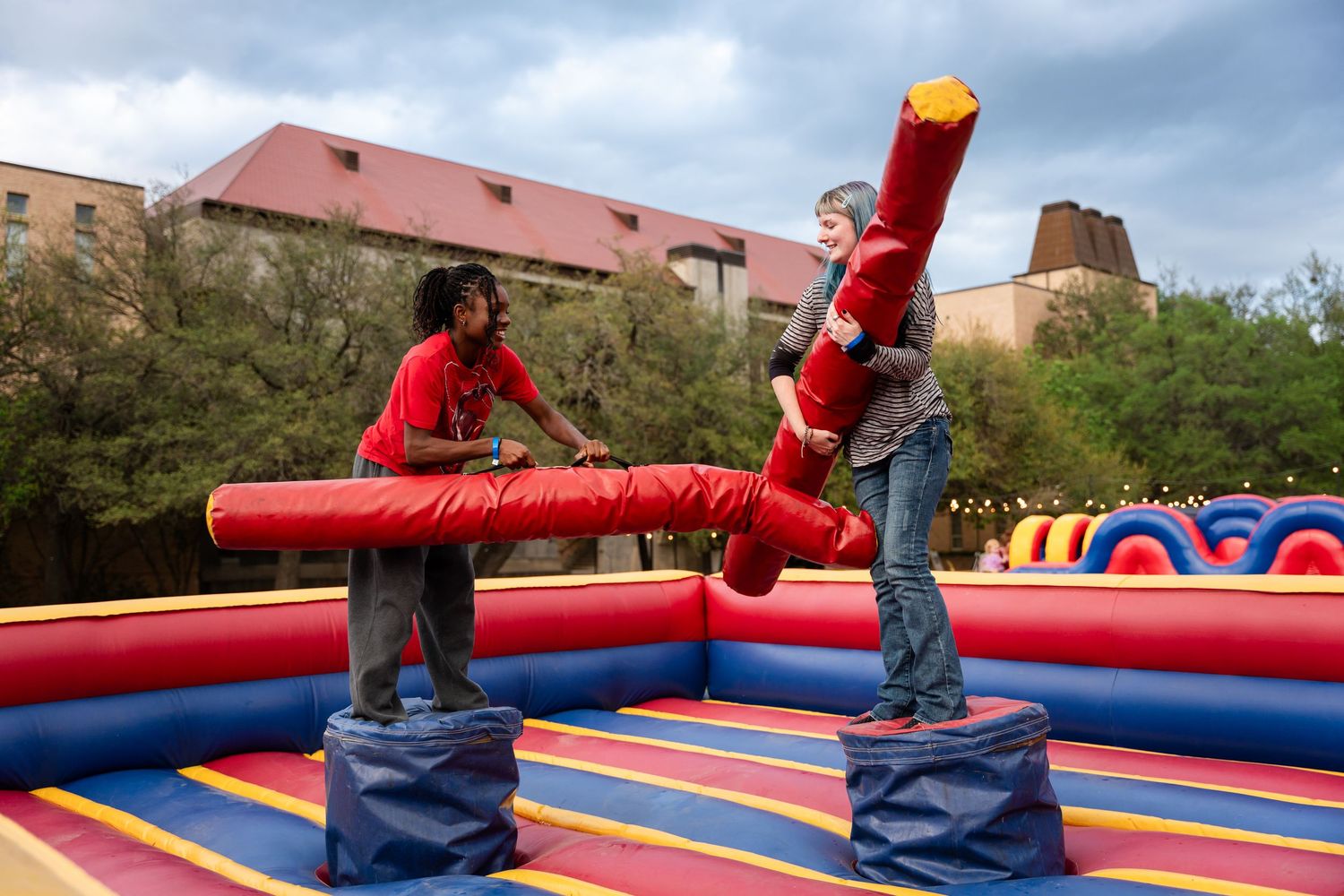 Two people compete in a jousting match on a colorful inflatable arena outdoors at a university event.