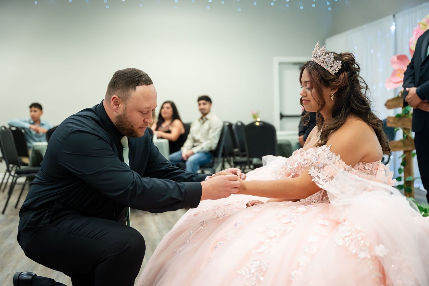 Man kneeling before a young woman in a pink quinceañera ball gown during a traditional ceremony indoors.
