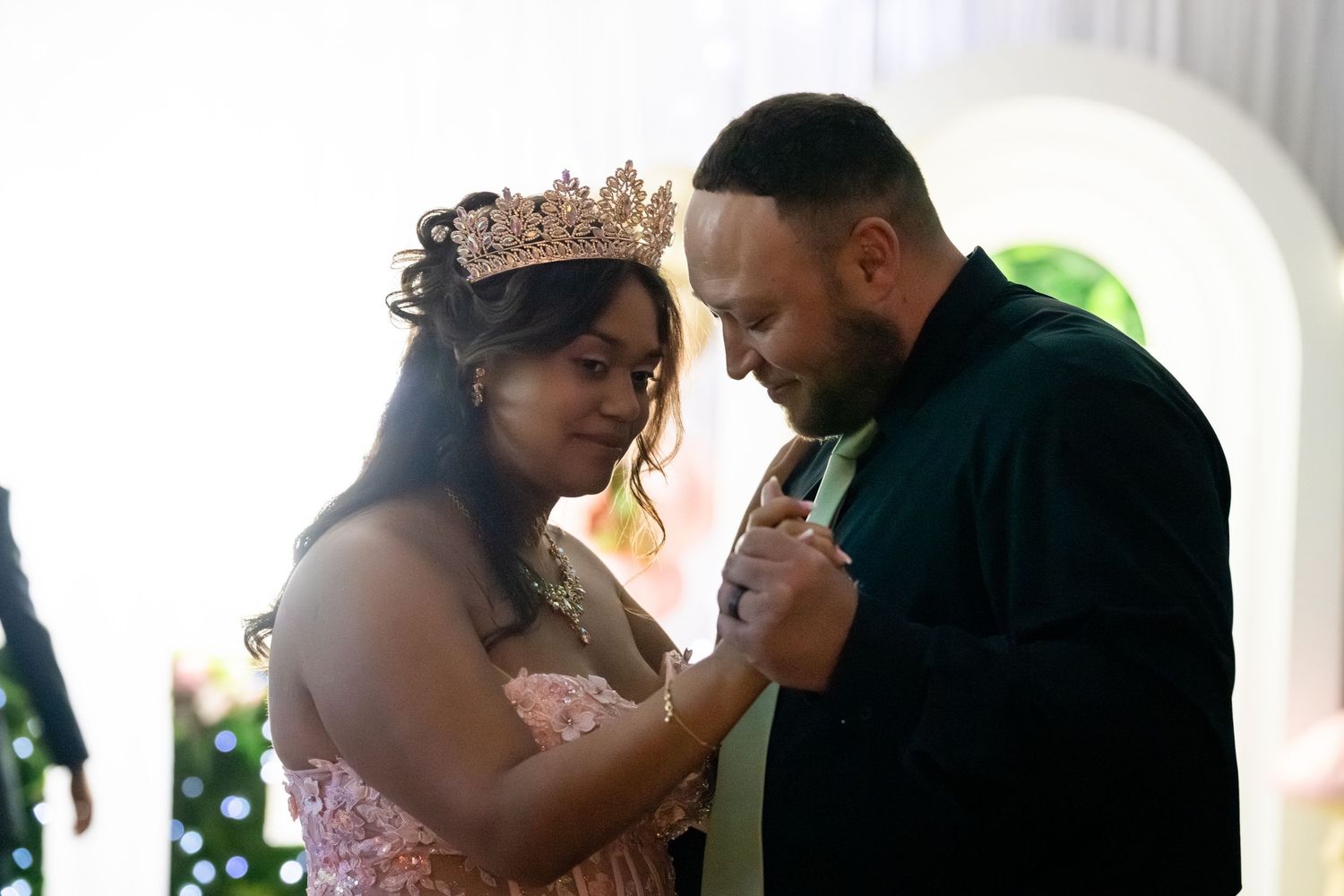 A woman wearing a rose gold crown and pink gown shares a tender dance moment with a man in a black shirt.