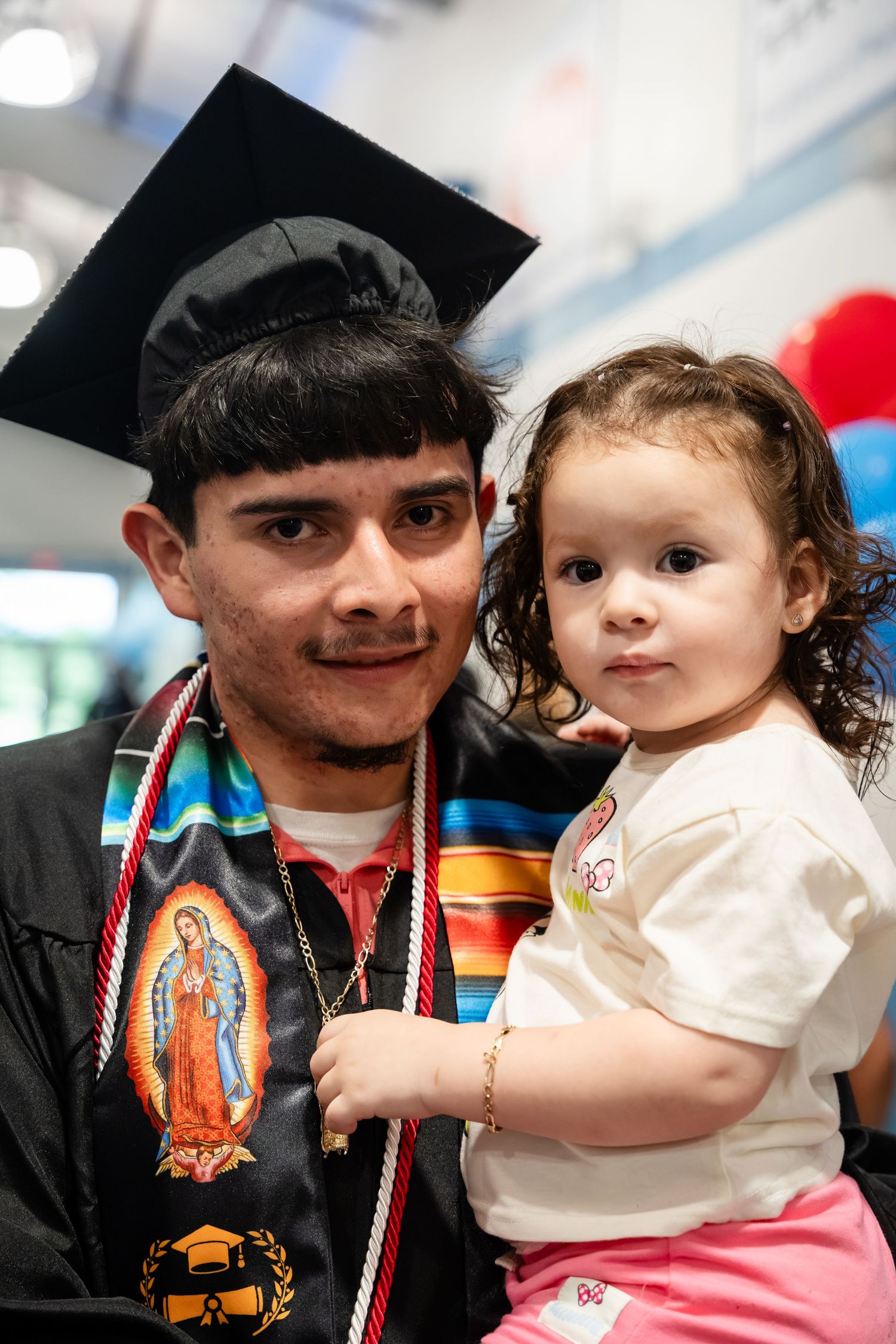 Graduate in cap and gown holds a young toddler at a graduation ceremony celebration.