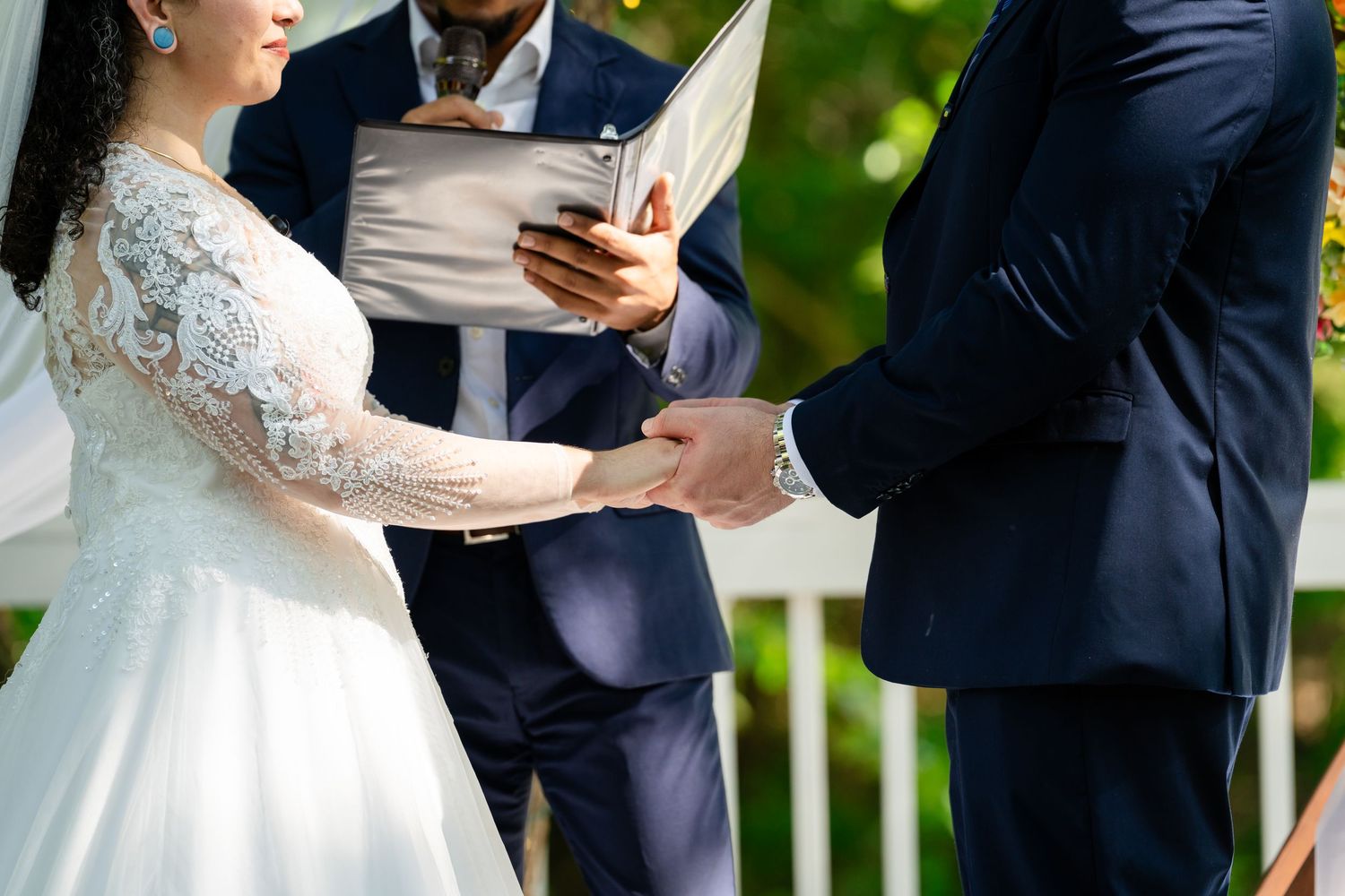 Bride and groom holding hands during outdoor wedding ceremony with officiant reading vows in background.