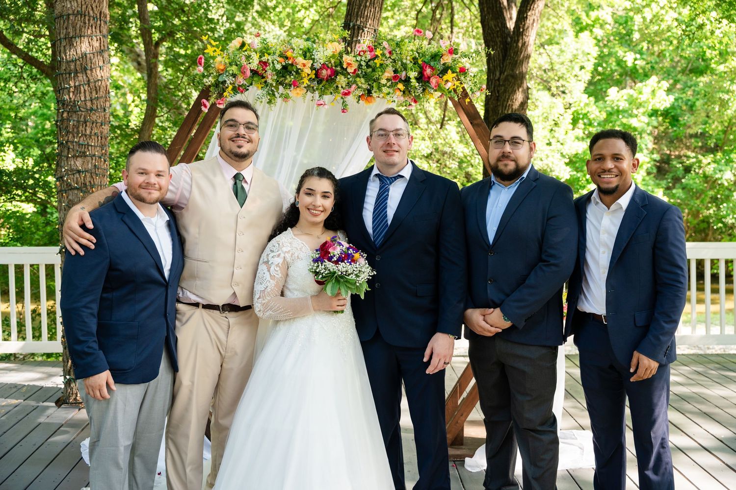 Bride and groom pose with four guests outdoors at a wedding ceremony beneath a floral arch on a wooden deck.