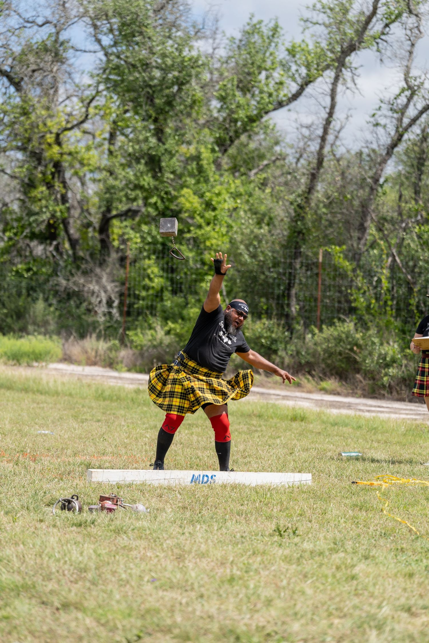 Man in yellow tartan kilt competing in Highland Games throwing event on grassy field with trees in background.