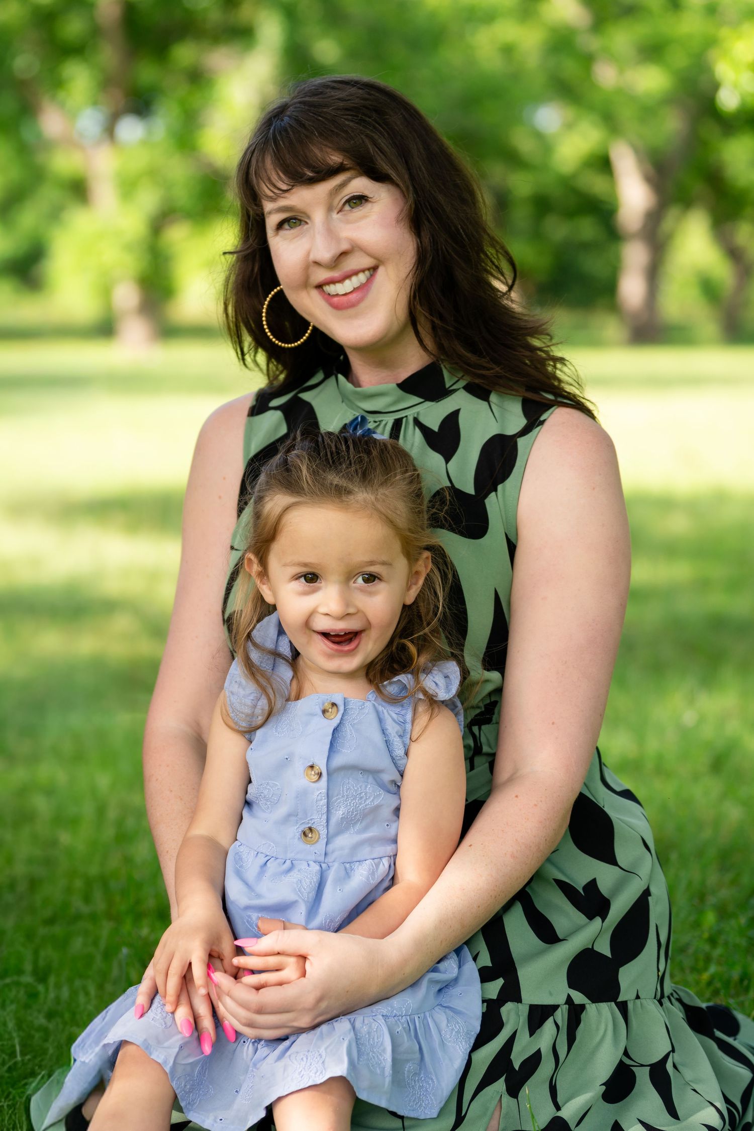 A smiling woman in a green floral dress sits outdoors holding a happy young girl in a blue dress in a park.