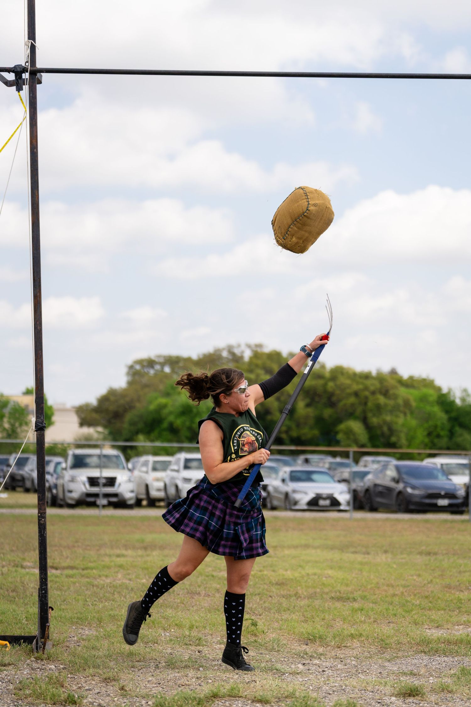 Woman in tartan skirt and boots throws weighted bag at Highland Games competition outdoors.