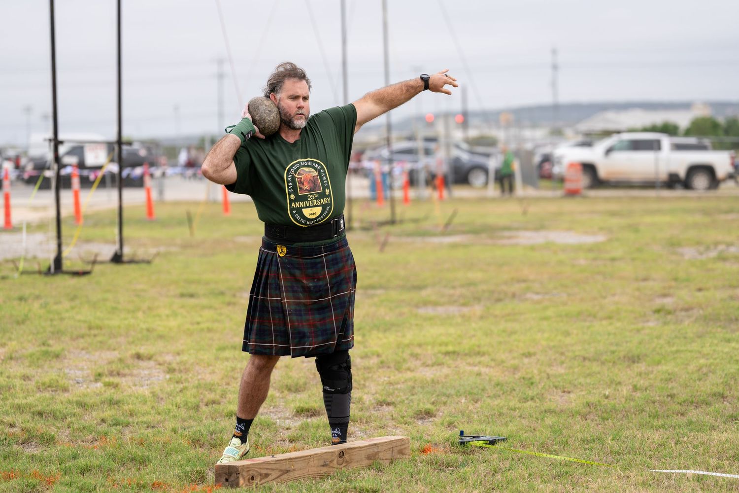 Man in tartan kilt throws shot put at Highland Games competition outdoors.